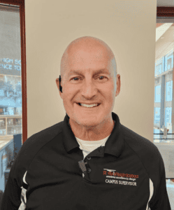 A smiling man in a black polo shirt with a campus supervisor label stands indoors, with a school background.